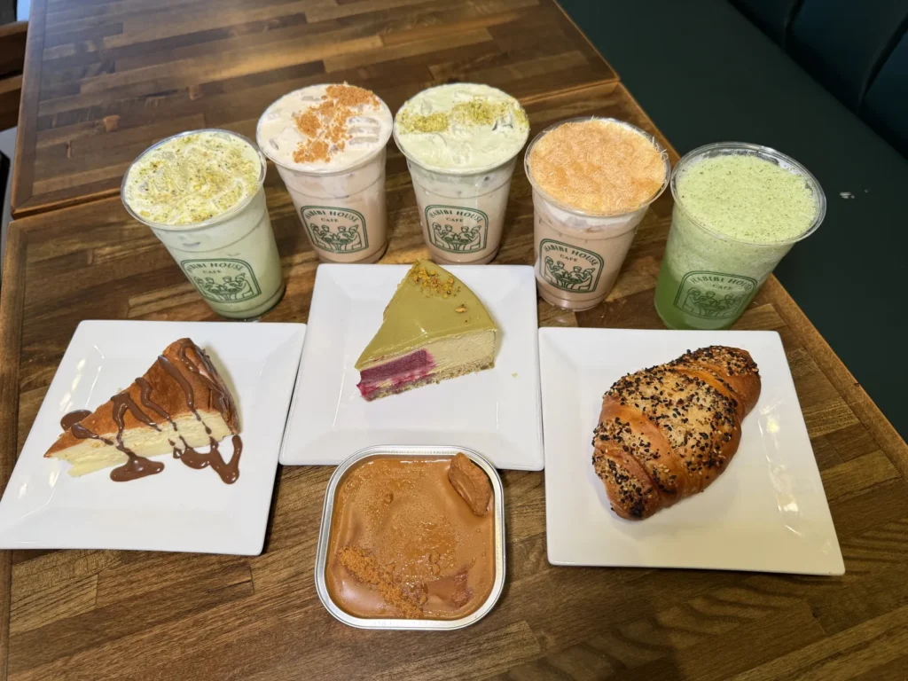 Five iced drinks in Habibi House Cafe cups beside a pistachio cheesecake slice, a chocolate-drizzled cheesecake, a spoonable tiramisu and a black-seed croissant