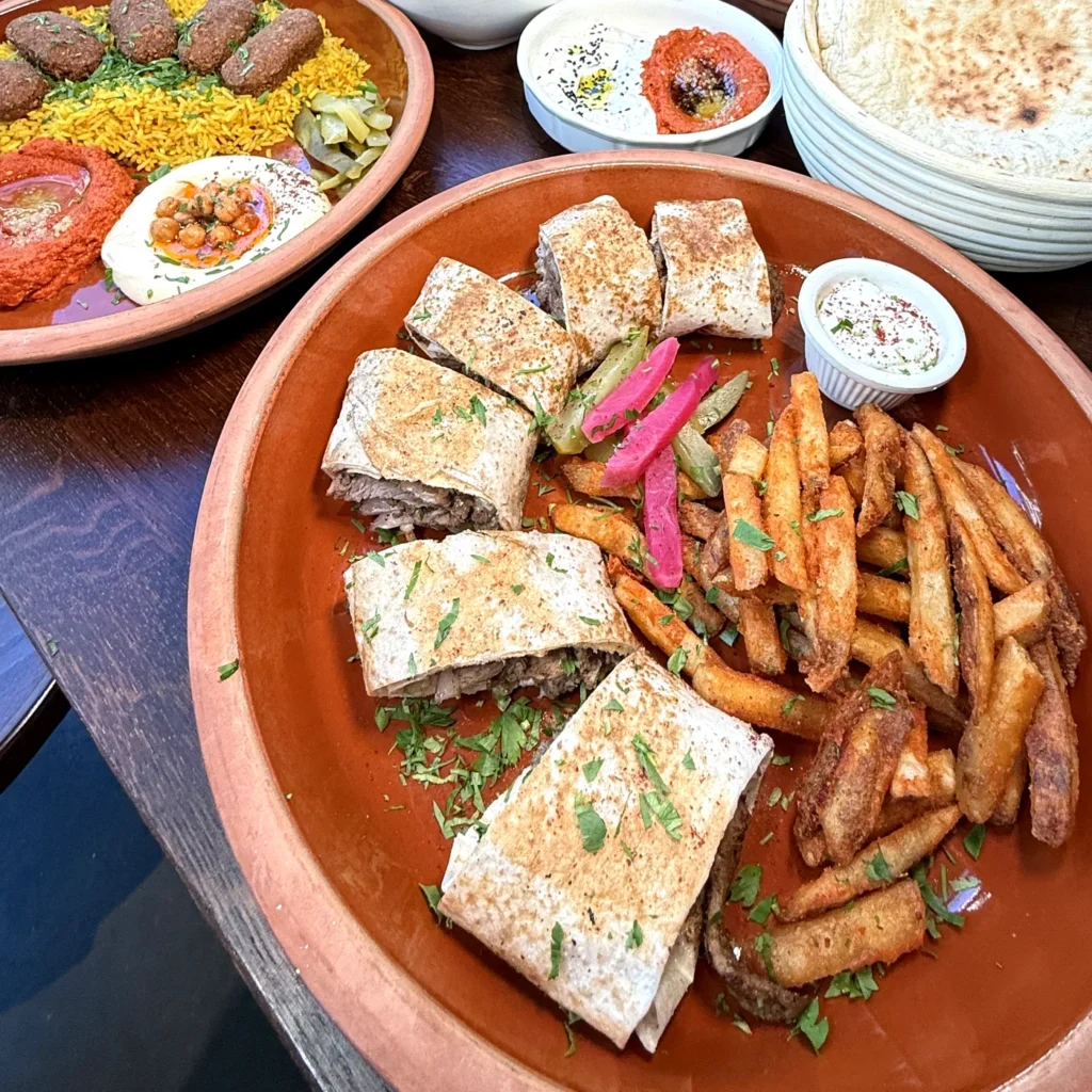 Araby shawarma sandwich cut into pieces with seasoned fries and garlic sauce at AYAT in Foggy Bottom, with a falafel platter behind it