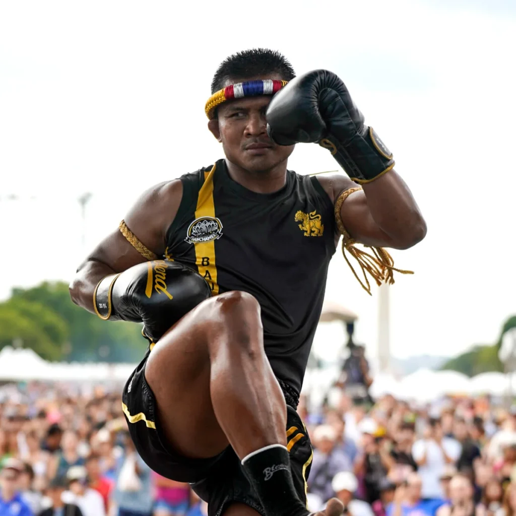 Buakaw Banchamek in black and gold Muay Thai shorts raising his knee mid-demonstration on stage, with the crowd behind him