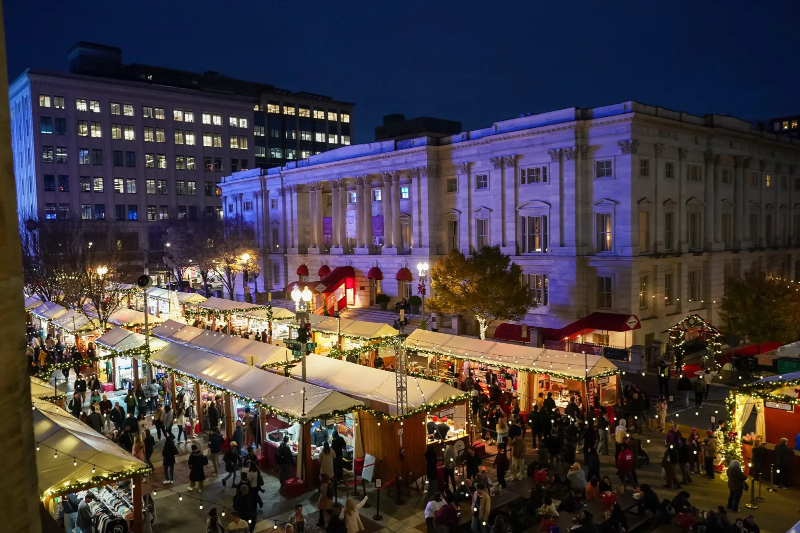 Rows of white vendor tents strung with lights fill F Street NW below the columns of the National Portrait Gallery at night during the DowntownDC Holiday Market