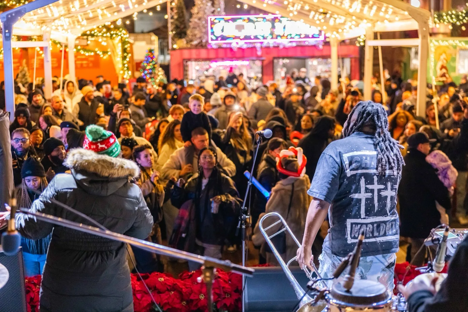 A band plays on the lit performance stage at the DowntownDC Holiday Market at night, seen from behind the musicians as a large crowd watches under string lights