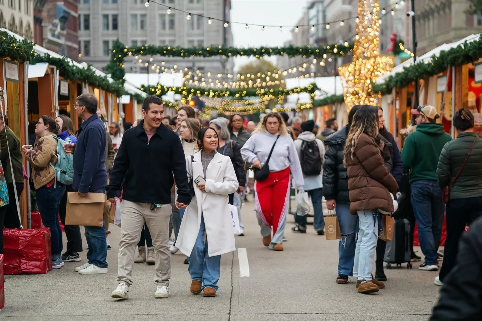 Shoppers carrying bags walk the center aisle between wooden vendor stalls under green garland and string lights on F Street NW during the day at the DowntownDC Holiday Market