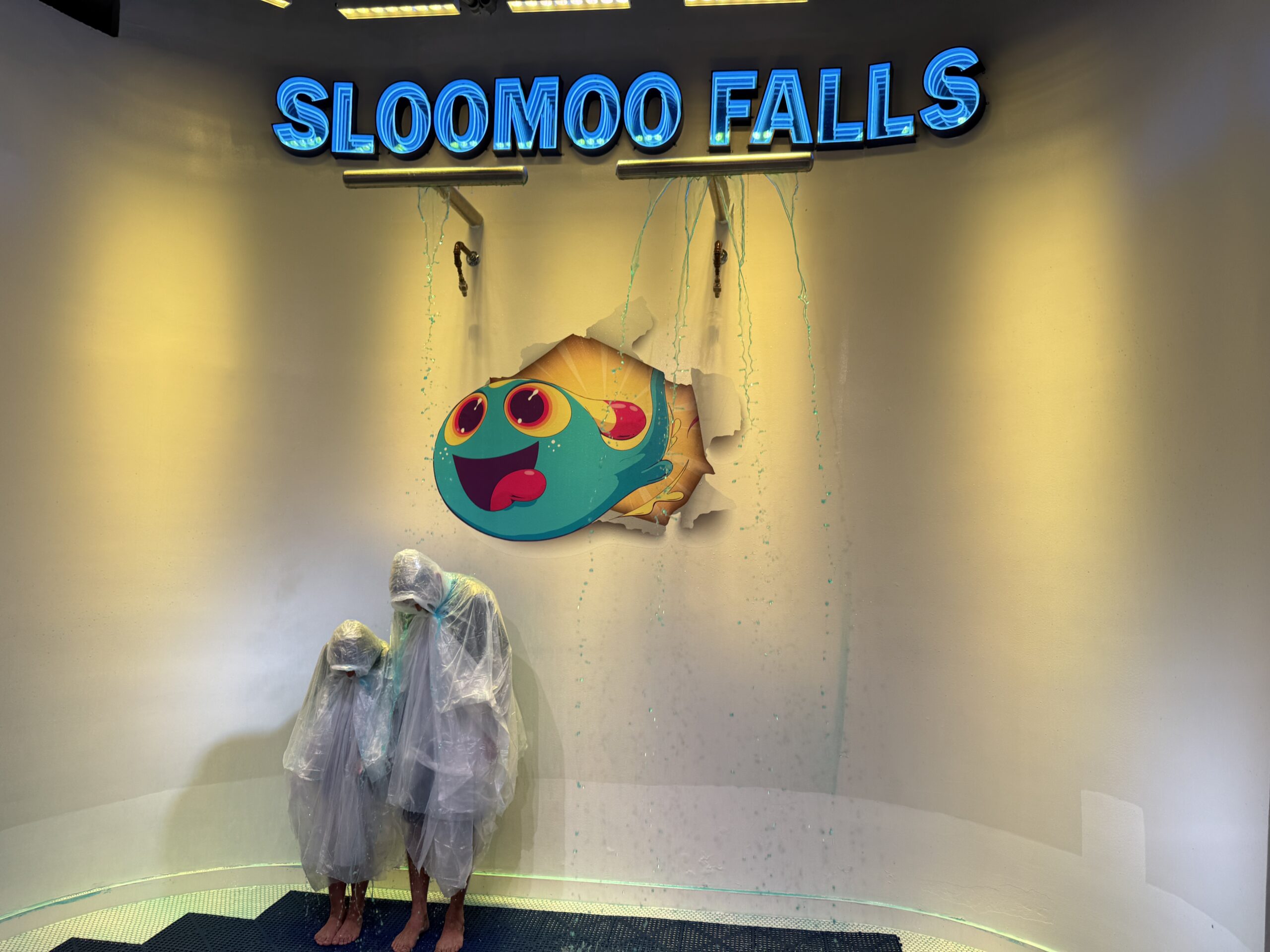 Two visitors wearing protective coverings beneath the Sloomoo Falls slime shower