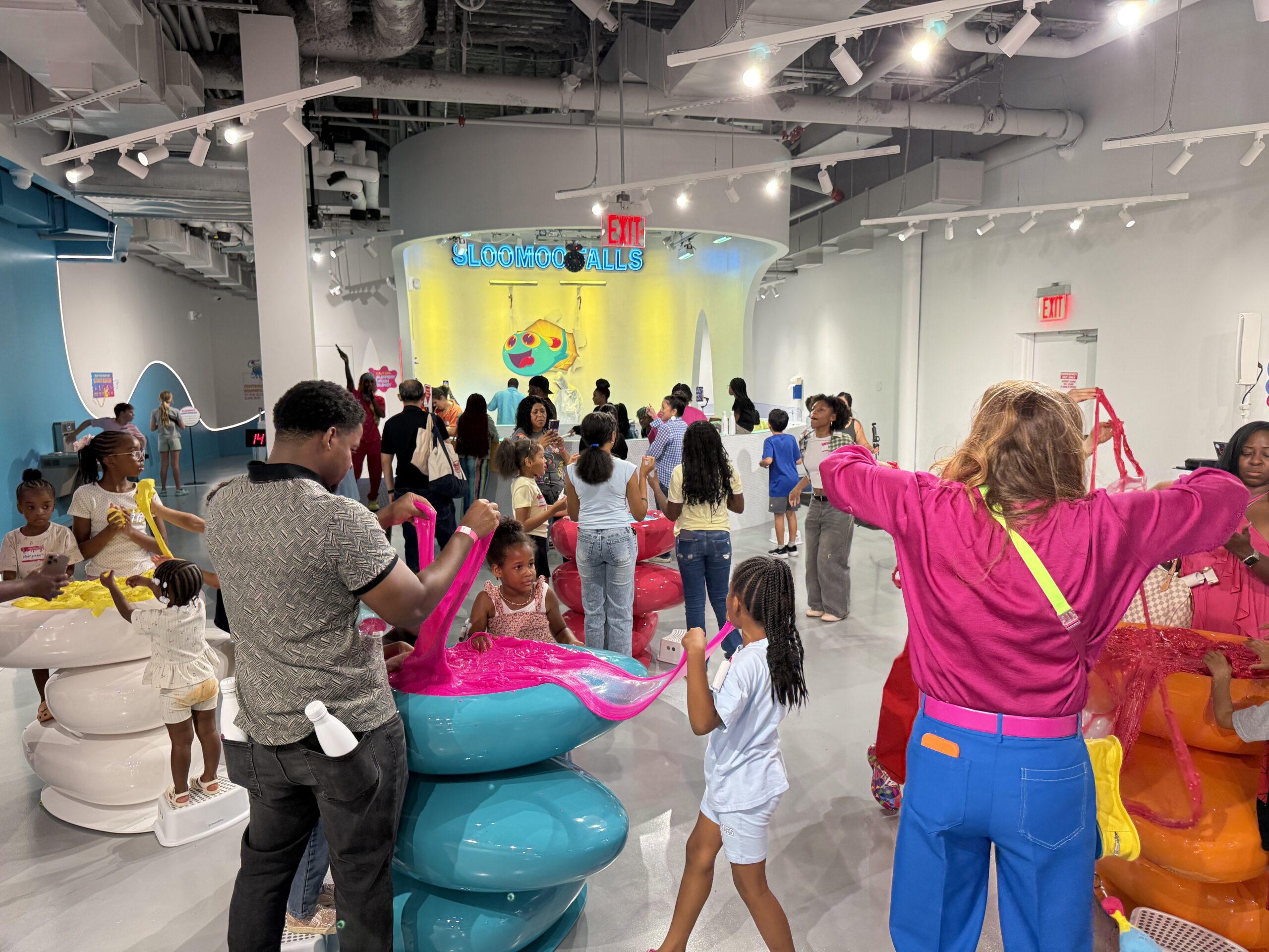 Visitors stretching colorful slime at Sloomoo Institute in Tysons Corner Center, with Sloomoo Falls in the background