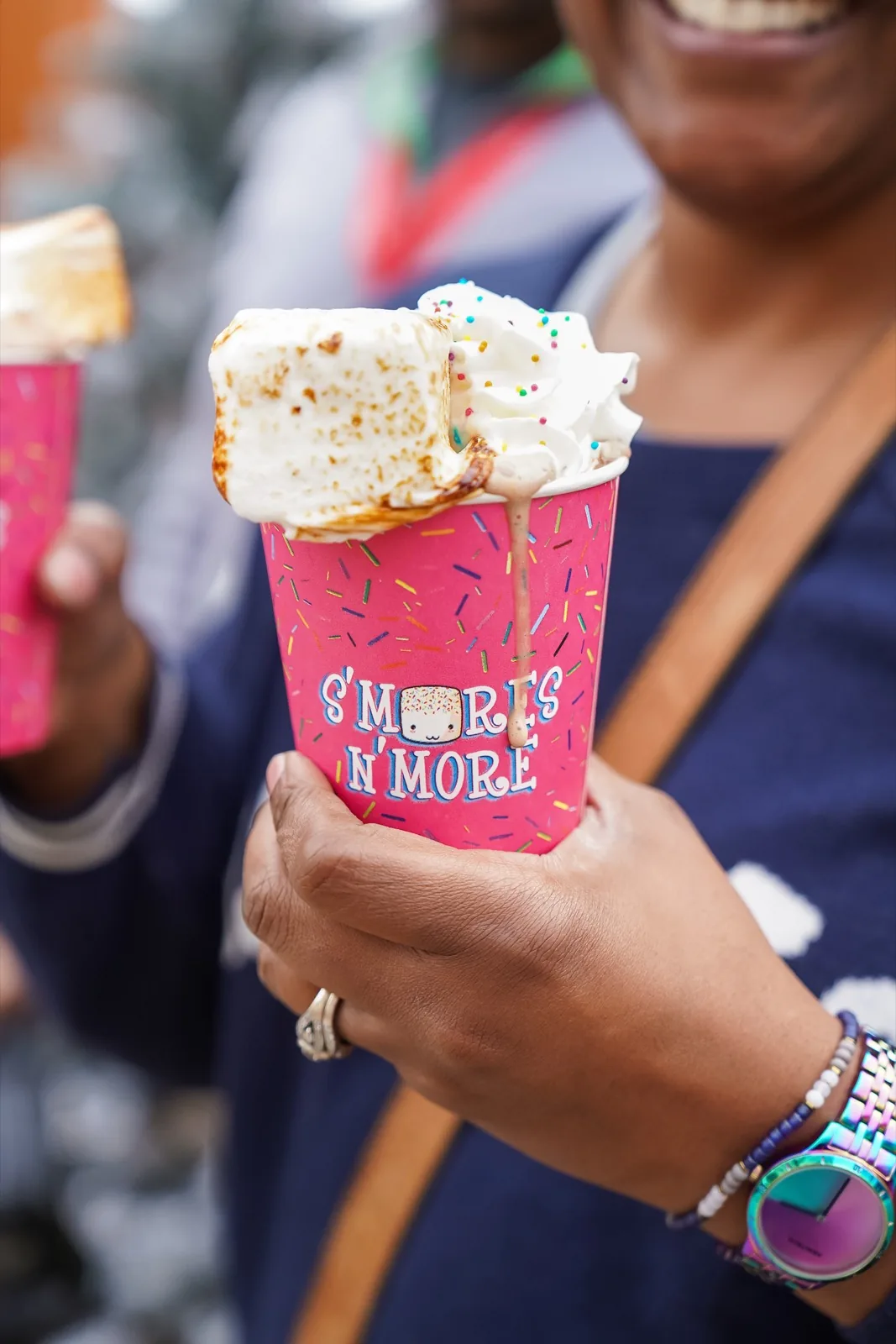 A hand holds a pink S’mores N’ More cup of hot chocolate topped with a torched marshmallow, whipped cream and sprinkles at the DowntownDC Holiday Market