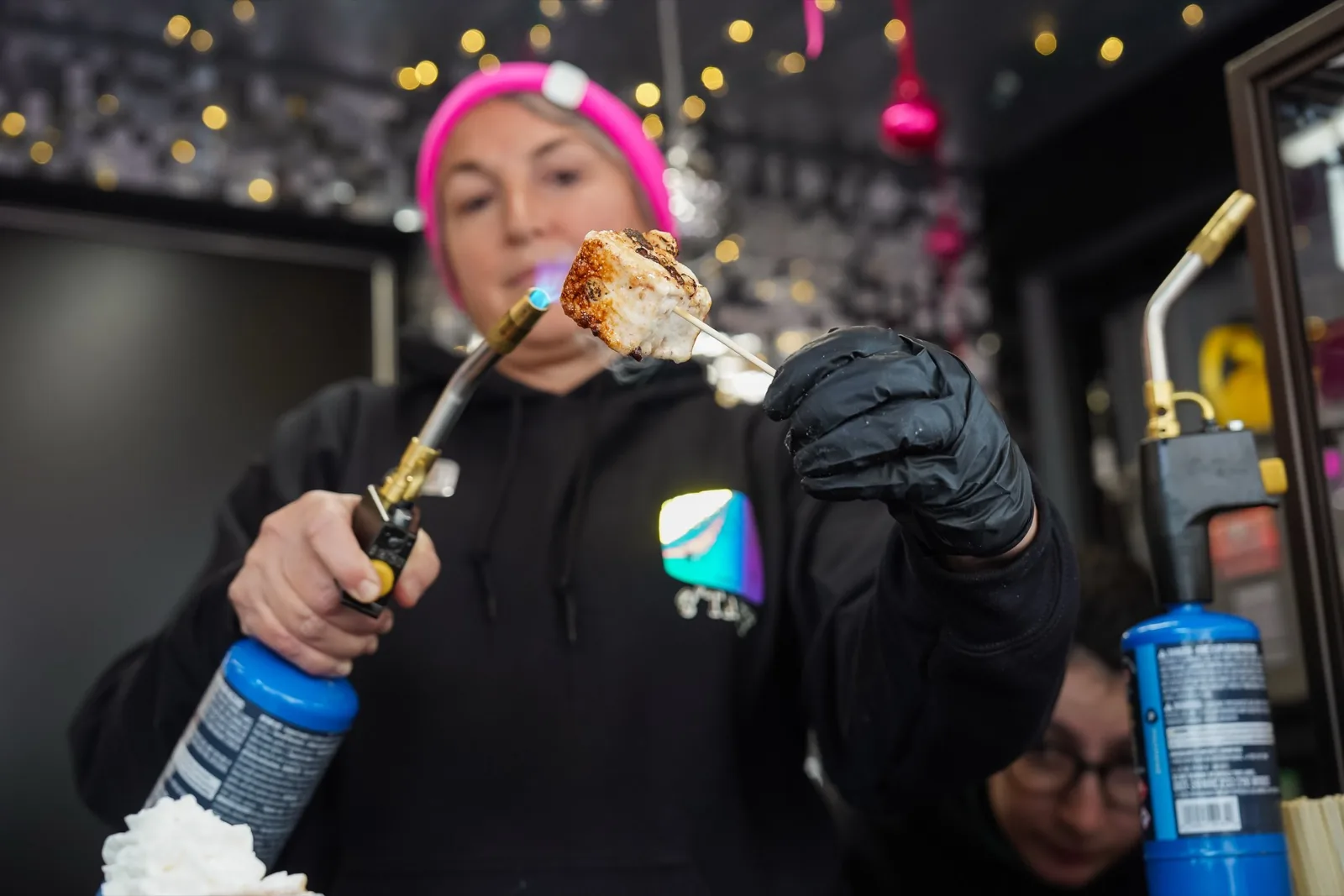 A S’mores N’ More vendor in a pink beanie torches a marshmallow on a stick with a handheld blowtorch inside the stall at the DowntownDC Holiday Market