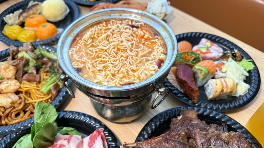 Plates from one lap of the UMI Alexandria buffet on a table: a personal hot pot of ramen, sliced beef, lamb chops, sushi, lo mein and dim sum
