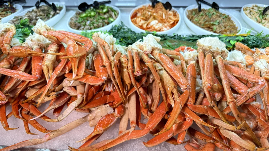 A pile of snow crab legs on ice at the UMI Alexandria seafood station, with kimchi and cold salads behind